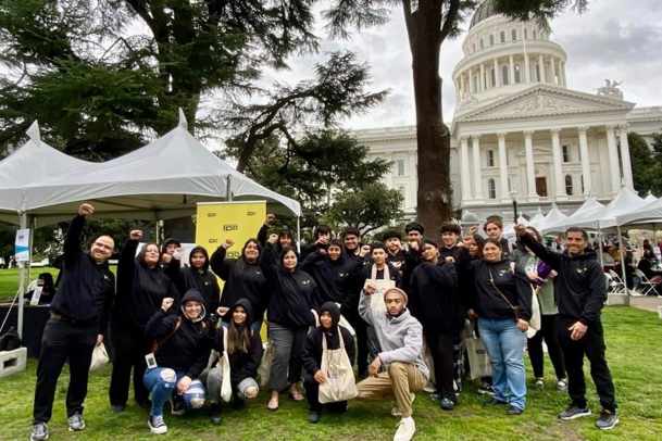 Pictured: A large group of participants pose together. Four young people are kneeling in front of others. Most are wearing black hoodies. They pose outside in front of tents with the State Capitol, a large white building, behind them in the distance. 