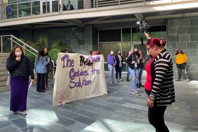 Pictured: Several individuals gather outside around a poster that says, 'The Problem Contains the Solution.' The word 'problem' is crossed out and says 'youth' slightly above it.  An individual in a striped sweater is in front of the poster holding a microphone.  