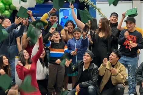 Pictured: A group of youth and young adults are gathered during a celebration. Everyone is dressed casually, wearing jeans and sneakers. Some individuals hold balloons and others throw green graduation caps in the air. They are indoors with a white and green balloon arch behind them. 