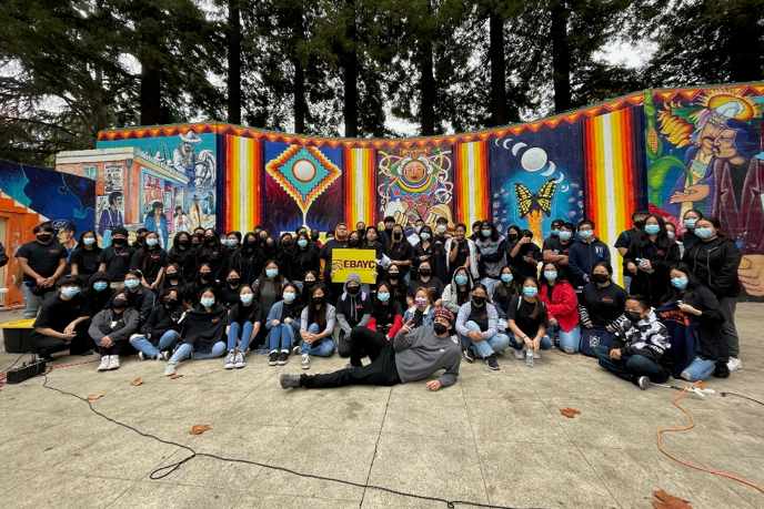 Pictured: A large group of youth stand and kneel together in front of a large colorful mural. They are outside at an event with multiple tall trees behind the mural in the distance. Most of the youth have a blue mask covering their nose and mouth. One youth hold a sign that says 'EBAYC.' 