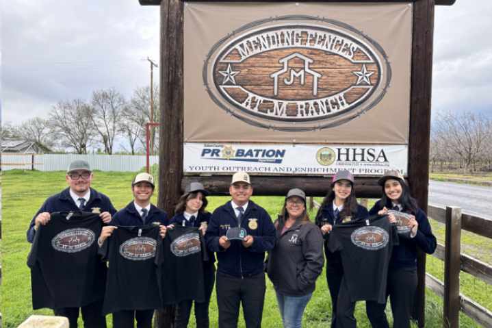 Pictured: Seven young smiling participants pose together. All wear back jackets and hats. Five participants are holding black shirts. They pose outside in front of a sign that says, 'Mending Fences at JM Ranch.'