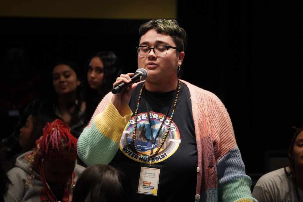 Pictured: A young participant is holding a microphone and wearing a black shirt and colorful cardigan. The photo is captured inside of a room and four individuals can be seen behind the participant