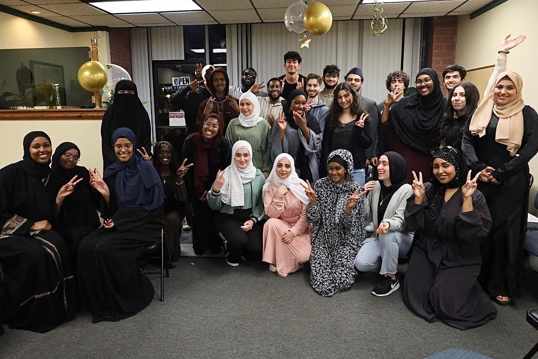 Pictured: A large group of 26 people pose together. Ten participants are kneeling in front of the individuals posing in the back. Many of the participants are wearing hijabs. They pose in a room with gold balloons hanging from the ceiling.