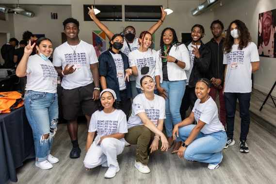 Pictured: 12 young participants pose together. Nine youth are standing in the back, three kneel in front of them. All wear white shirts with black writing that says, 'Kickback With The Youth.' They pose in a room with colorful artwork.