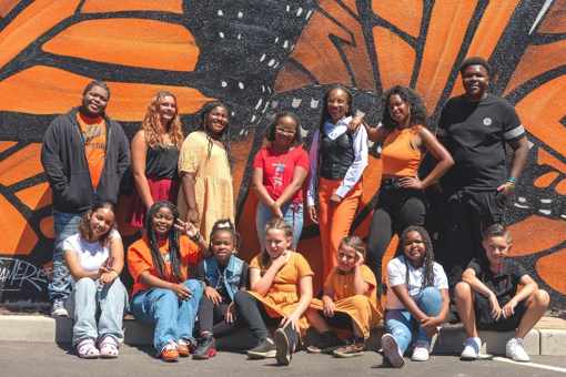 Pictured: 14 young participants pose together. Seven young people are standing in the back, seven kneel in front of them. They pose outside in front of a mural of a large orange butterfly.